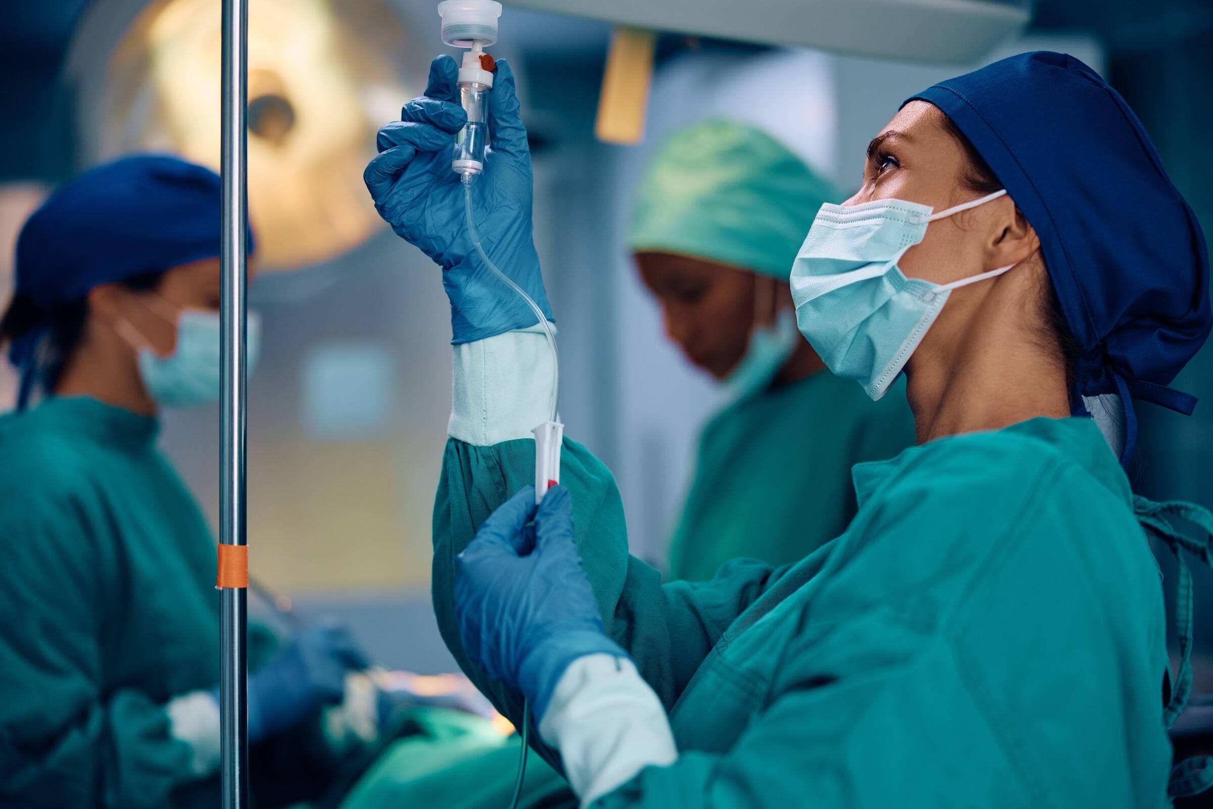 A nurse in a surgical cap and mask adjusting an intravenous line in an operating room